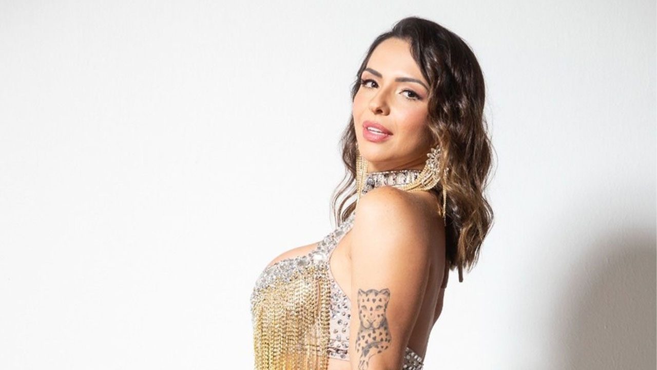 Karol Rosalin posa de lado com top bordado e franjas douradas, olhando por cima do ombro para a câmera, com tatuagem de onça no braço