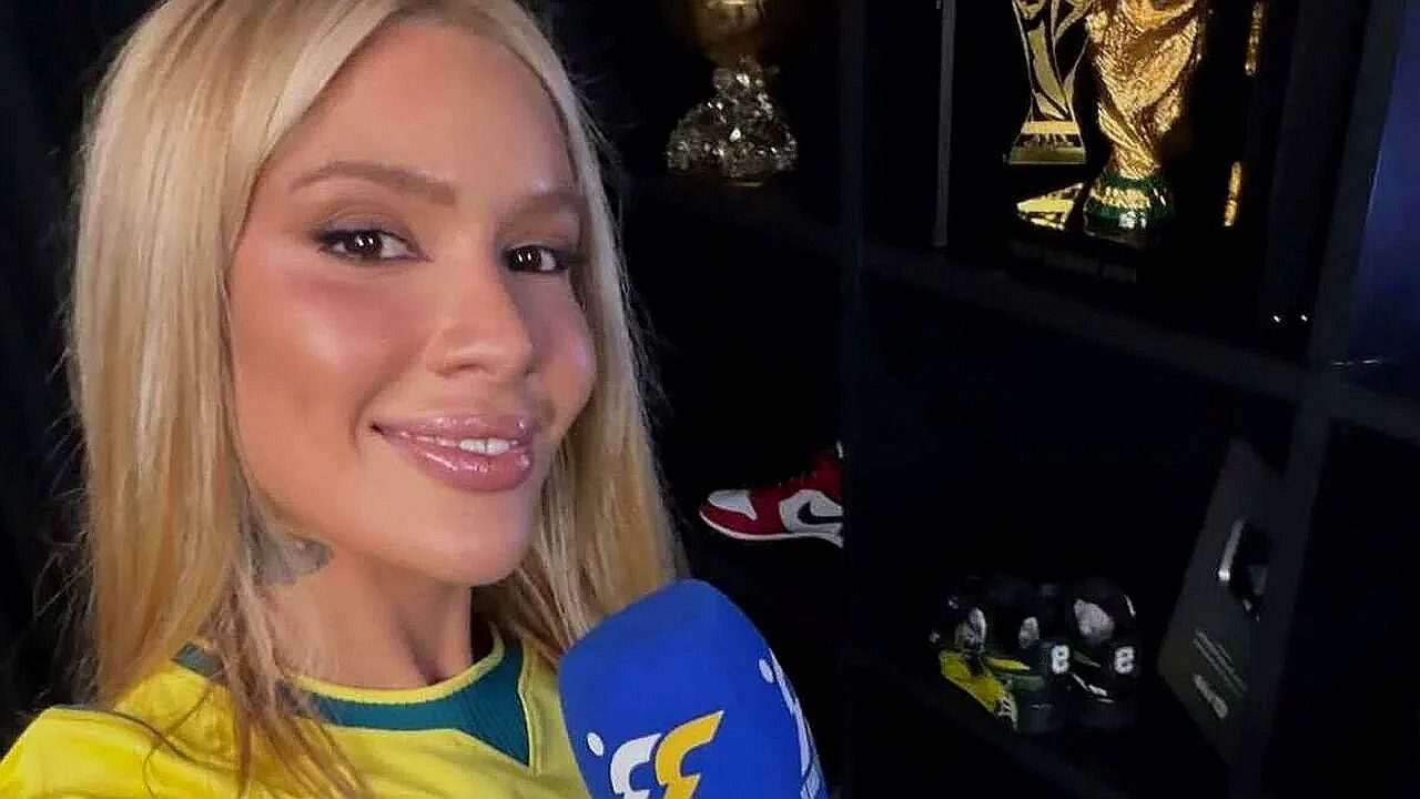 Mulher com camisa do Brasil segura microfone em gravação com prateleira ao fundo exibindo troféu da Copa do Mundo