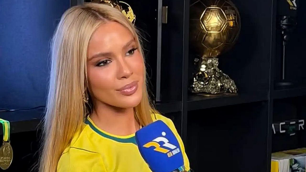 Mulher loira com camisa da seleção brasileira concede entrevista diante de prateleira com troféus