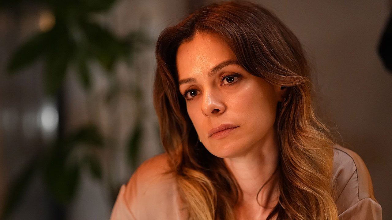 Özge Özpirinçci como Karsu na novela turca Coração de Mãe com expressão serena, cabelos castanhos ondulados e blusa de tom neutro em cenário doméstico iluminado