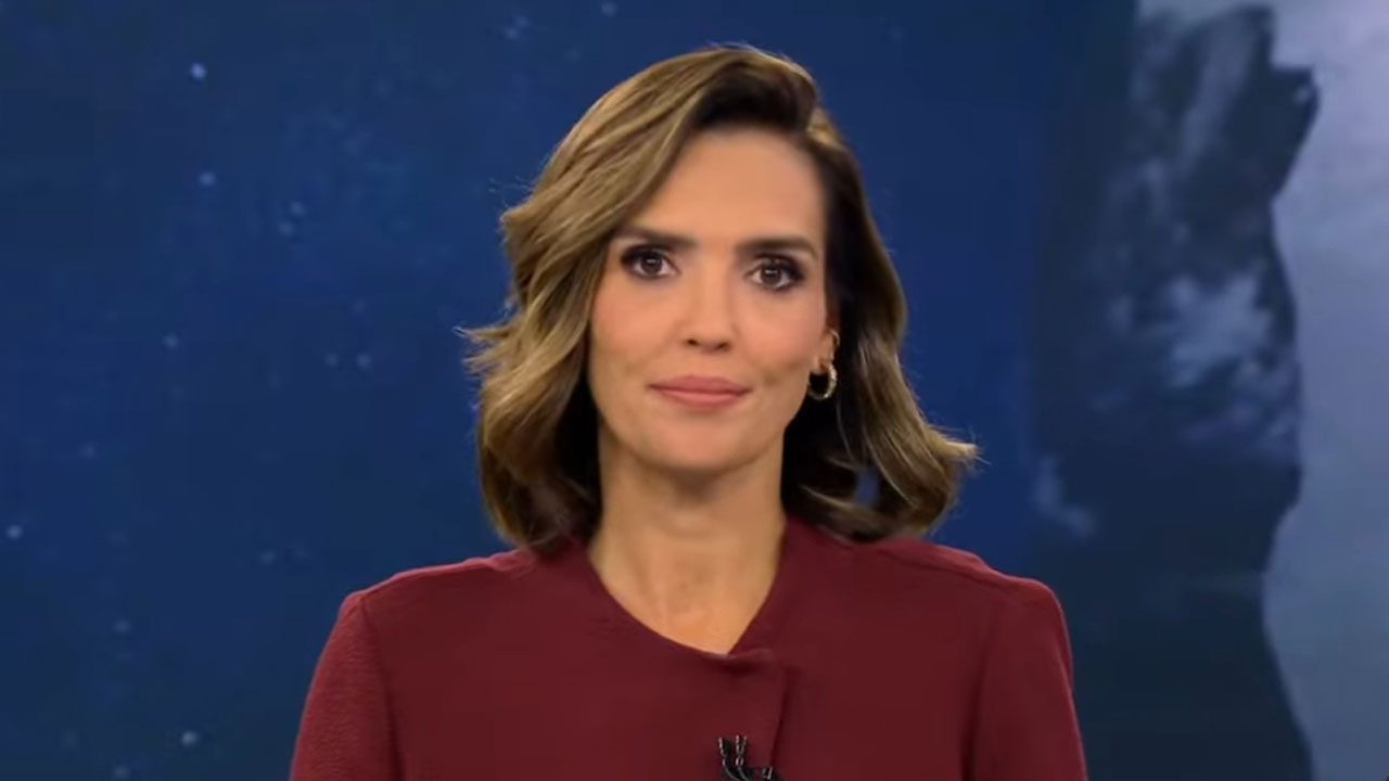 Karyn Bravo com blazer vinho e cabelos ondulados em plano médio durante a apresentação do Jornal da Cultura