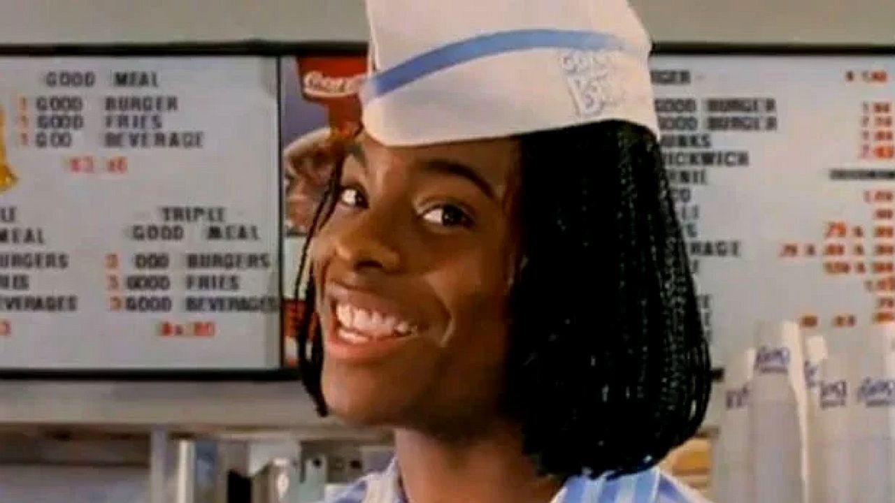 Funcionário de lanchonete sorri usando chapéu branco diante de cardápio com preços de hambúrgueres e combos ao fundo