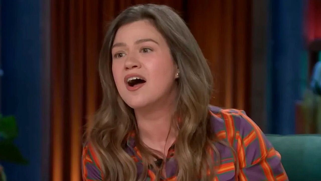 Mulher jovem com cabelos castanhos fala animadamente em um programa de televisão, usando camisa colorida listrada, com expressão de surpresa ou entusiasmo enquanto olha para alguém fora da câmera.