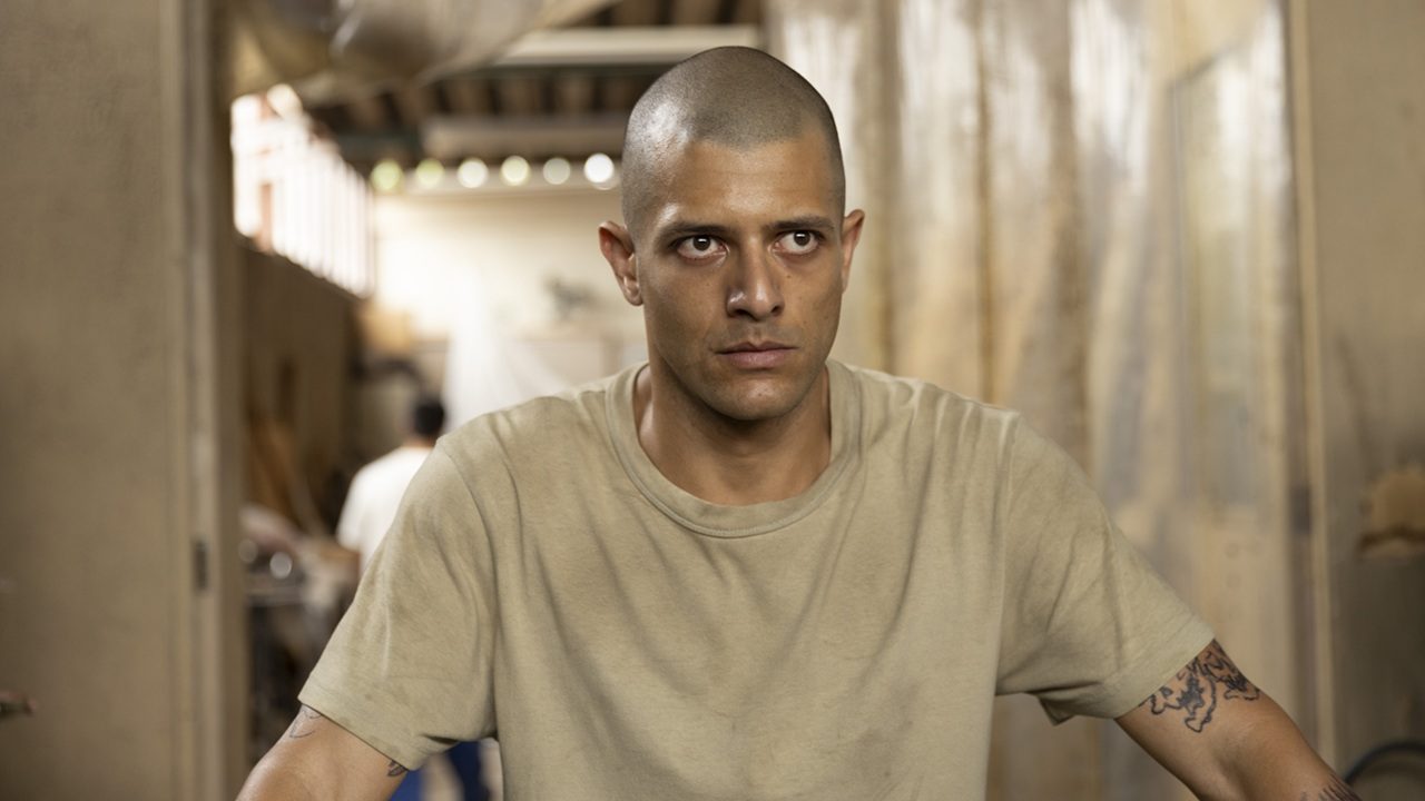 Homem de cabeça raspada usa camiseta bege e encara à frente com expressão tensa em corredor estreito de ambiente desgastado