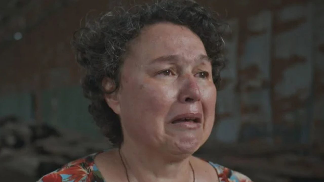 Mulher de meia-idade com cabelos curtos e cacheados aparece chorando, com expressão de dor e sofrimento. Seu rosto está avermelhado e molhado por lágrimas. Ela veste uma blusa estampada com flores e está em um ambiente interno escuro e desgastado ao fundo