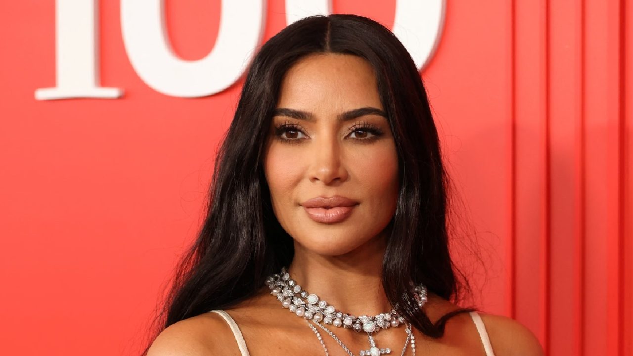 Kim Kardashian em evento de gala, com vestido claro de alças finas, colares de diamantes e cabelo solto, posando diante de um fundo vermelho
