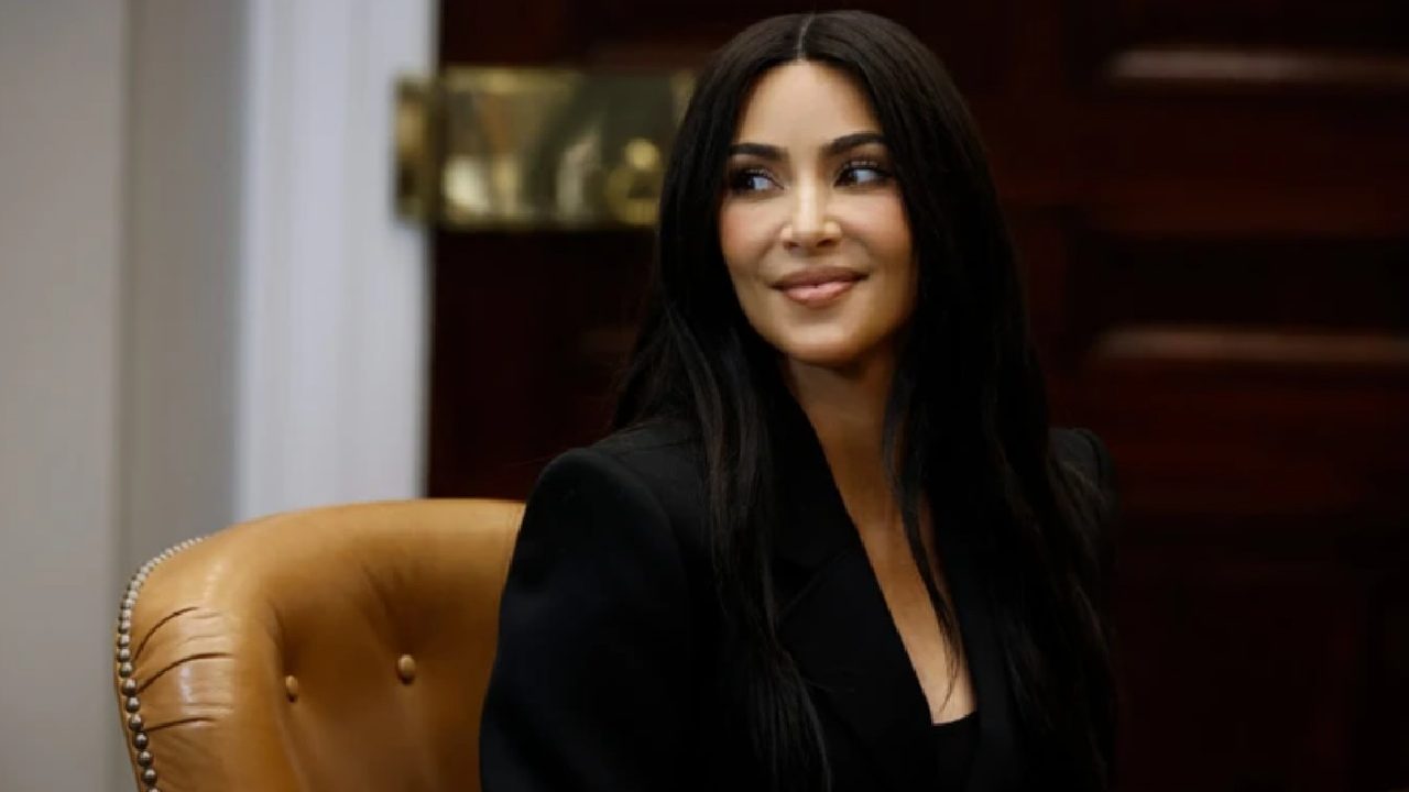 Mulher de cabelos longos e escuros, vestindo blazer preto, sorri enquanto senta em poltrona de couro em ambiente formal.