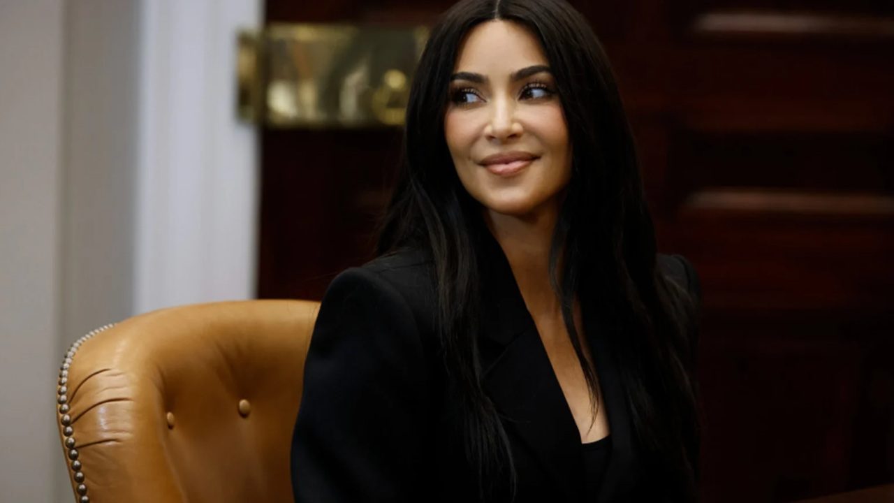 A imagem mostra uma mulher sentada em uma cadeira de couro marrom, usando um blazer preto e com os cabelos longos e escuros soltos. Ela sorri levemente enquanto olha para o lado, em um ambiente que parece ser um escritório ou sala de reuniões, com móveis de madeira ao fundo