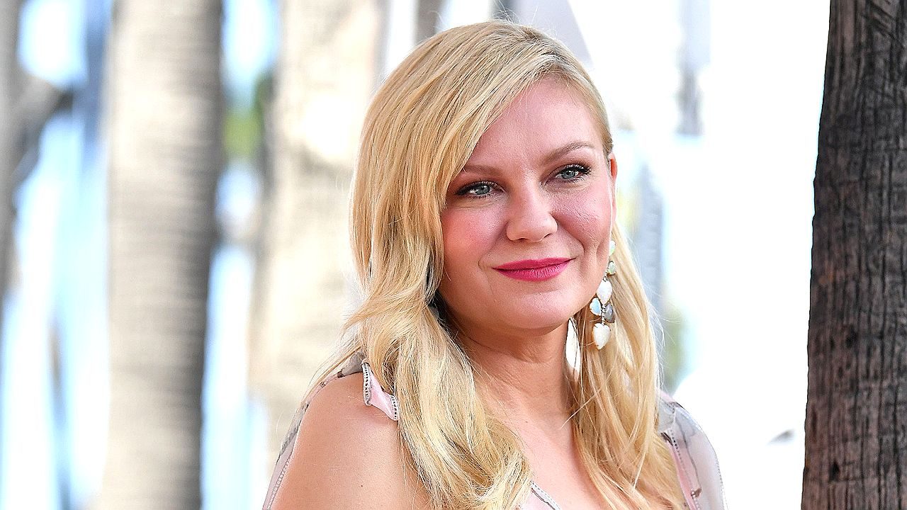 Atriz loira sorri ao ar livre com árvores ao fundo em evento ou entrevista informal