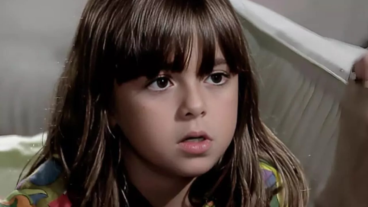 Klara Castanho com expressão séria enquanto está sentada usando roupa colorida em cena de produção televisiva brasileira