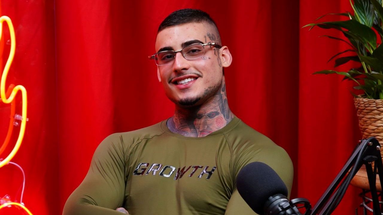 Kylian Cria sorri com óculos e braços cruzados vestindo camiseta verde da Growth durante gravação de podcast em estúdio com fundo vermelho