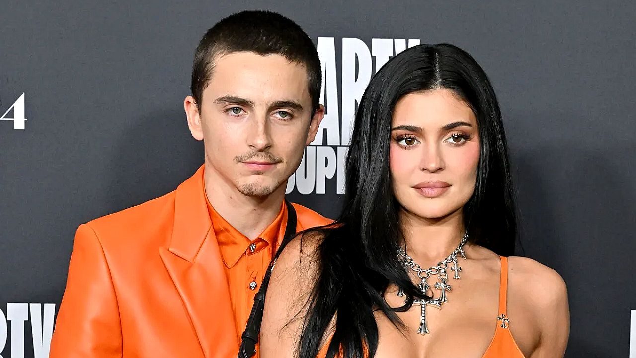 Homem e mulher posam juntos em evento com roupas laranja diante de painel com logotipo de estúdio