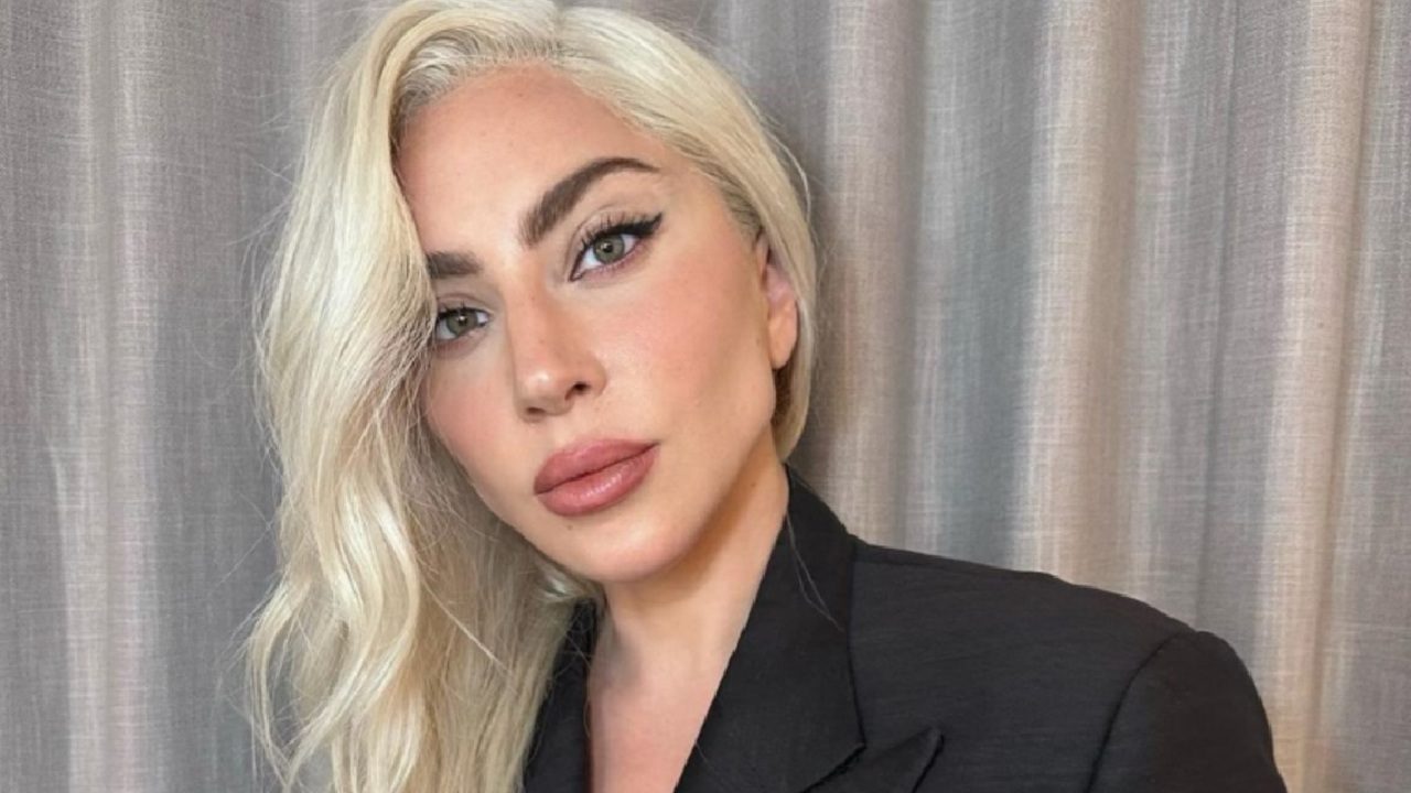 A imagem mostra uma mulher de cabelos loiros platinados e maquiagem elegante, com batom rosado e delineado marcado nos olhos. Ela veste um blazer preto e posa diante de uma cortina cinza, em um ambiente interno bem iluminado