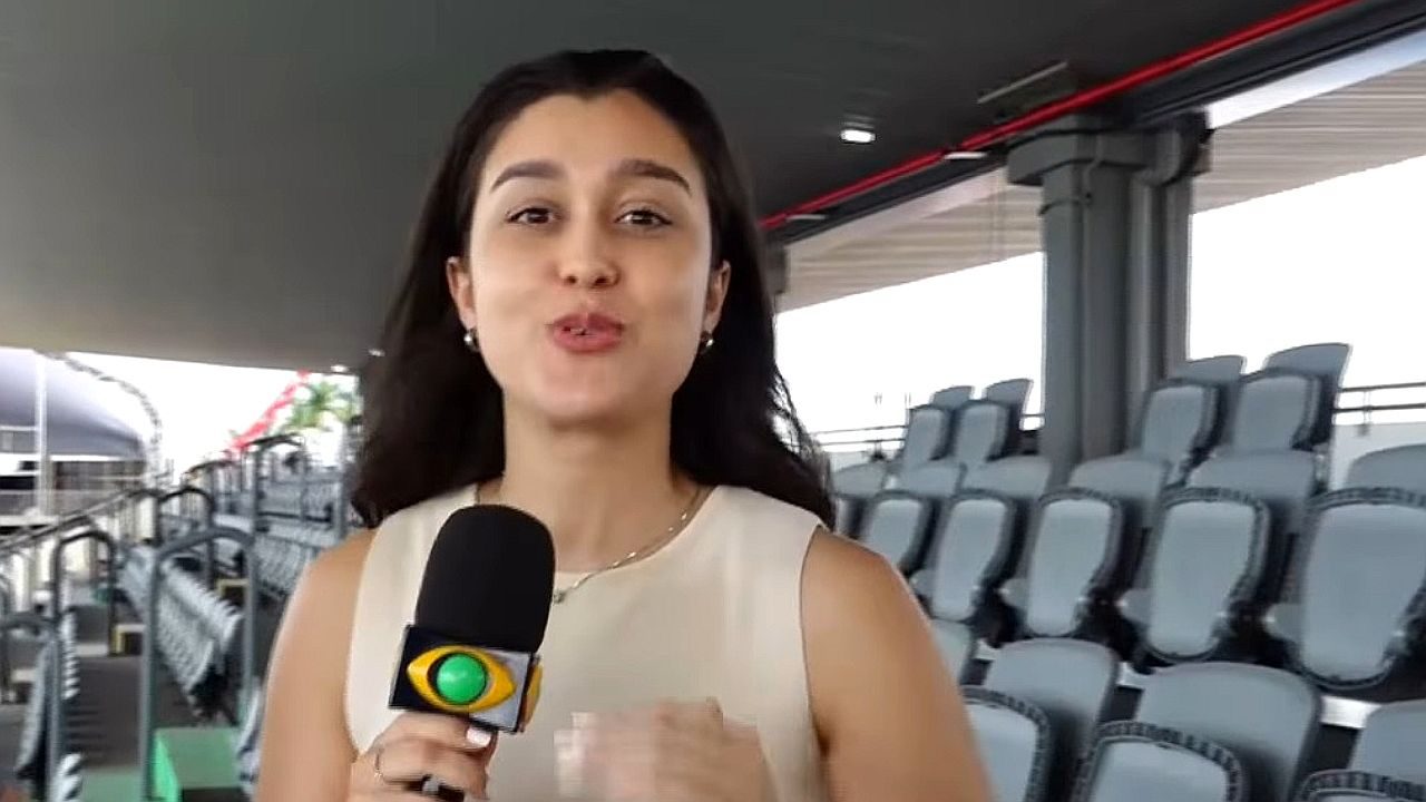 Uma repórter está em um ambiente que parece ser uma arquibancada de estádio ou arena, com várias fileiras de assentos ao fundo. Ela segura um microfone com logotipo de emissora e fala diretamente para a câmera. Usa uma blusa clara sem mangas e tem os cabelos longos soltos. A estrutura do local inclui cobertura, colunas e corrimãos, sugerindo um espaço para eventos ou transmissões esportivas.