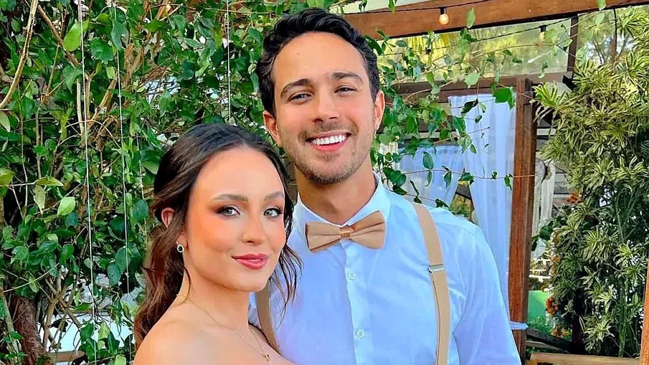 Casal sorridente posa junto em ambiente externo com plantas ao fundo em foto de evento social