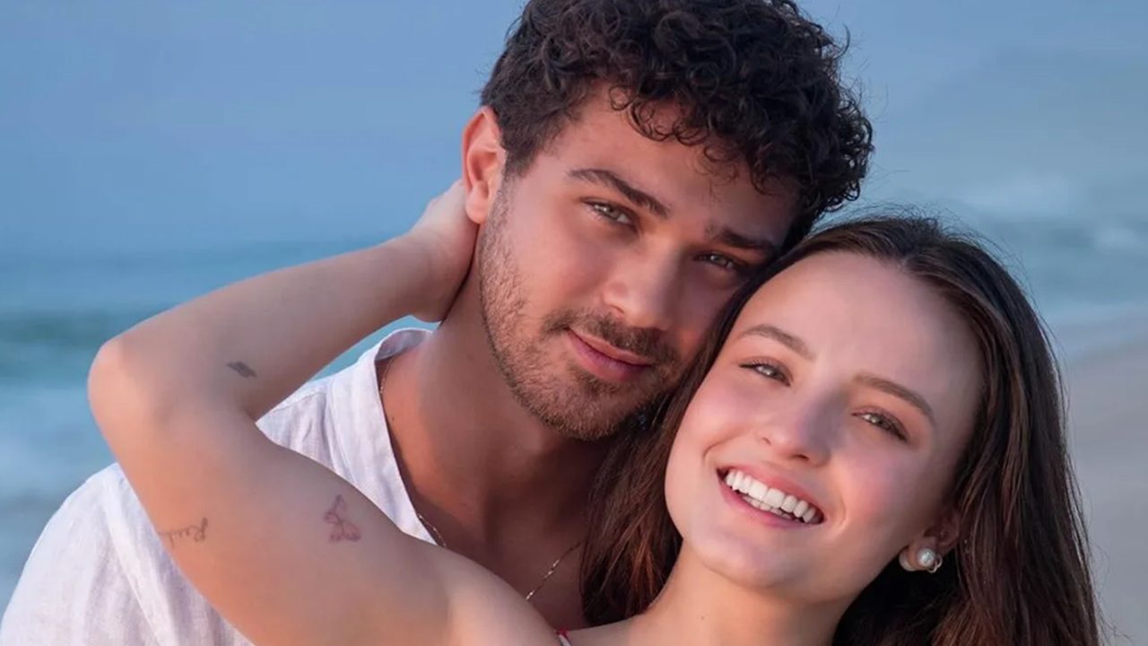A imagem mostra um casal sorridente em um ambiente de praia. Eles estão próximos um do outro, com o mar e o céu azul ao fundo. A mulher abraça o homem pelo pescoço, e ambos parecem felizes e relaxados, transmitindo um clima romântico e natural