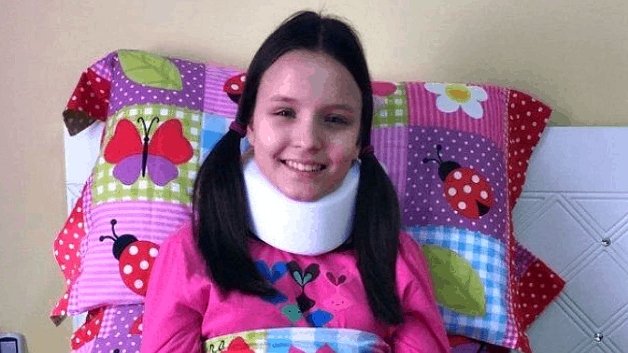 Menina sorri usando colar cervical enquanto está deitada em cama com almofada colorida