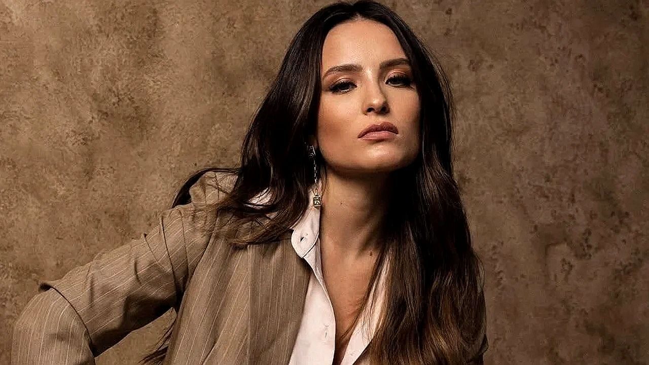 Larissa Manoela usa blazer bege e camisa branca em ensaio fotográfico com expressão séria diante de fundo marrom