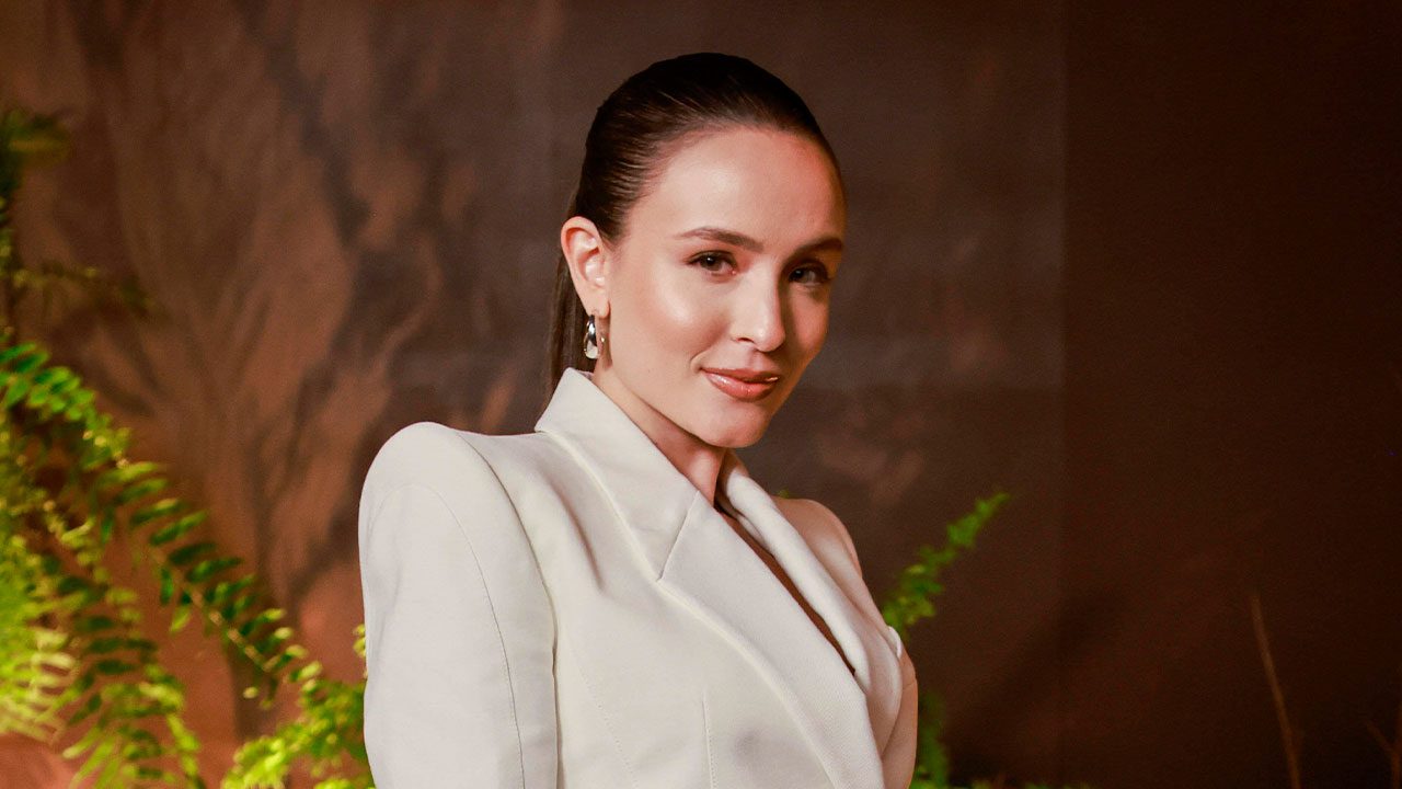 Atriz Larissa Manoela com cabelo preso e maquiagem natural, usando blazer branco em um evento com plantas no fundo