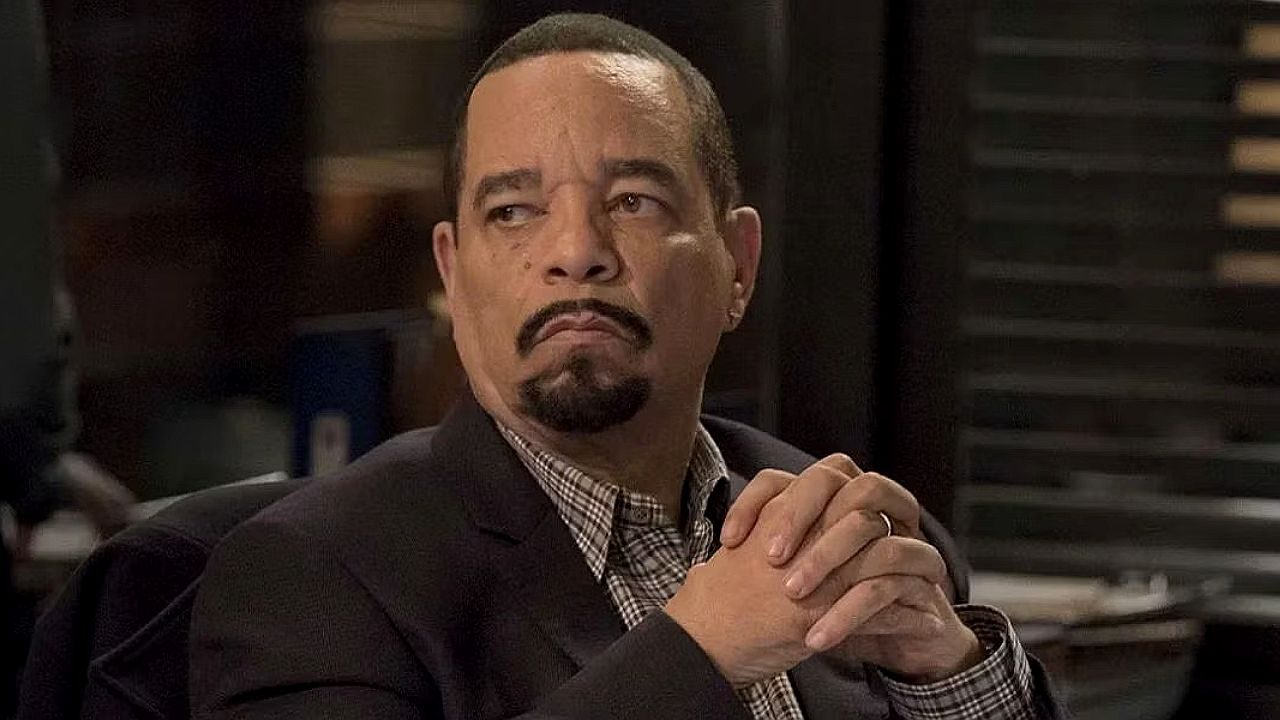 Homem de terno com mãos entrelaçadas observa ao lado em cena de série policial ambientada em escritório