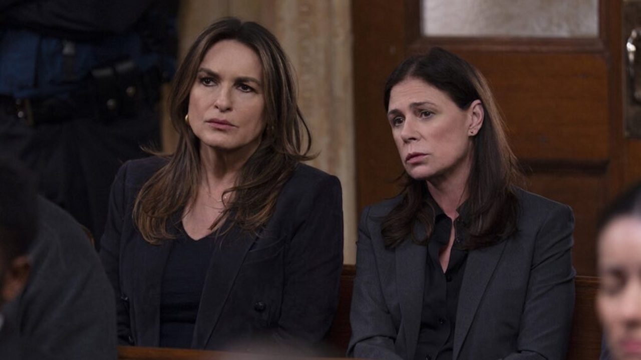 Duas mulheres sentadas lado a lado em um tribunal observam algo com expressões sérias, ambas vestindo ternos escuros, com fundo mostrando madeira, parede bege e outras pessoas sentadas ao redor