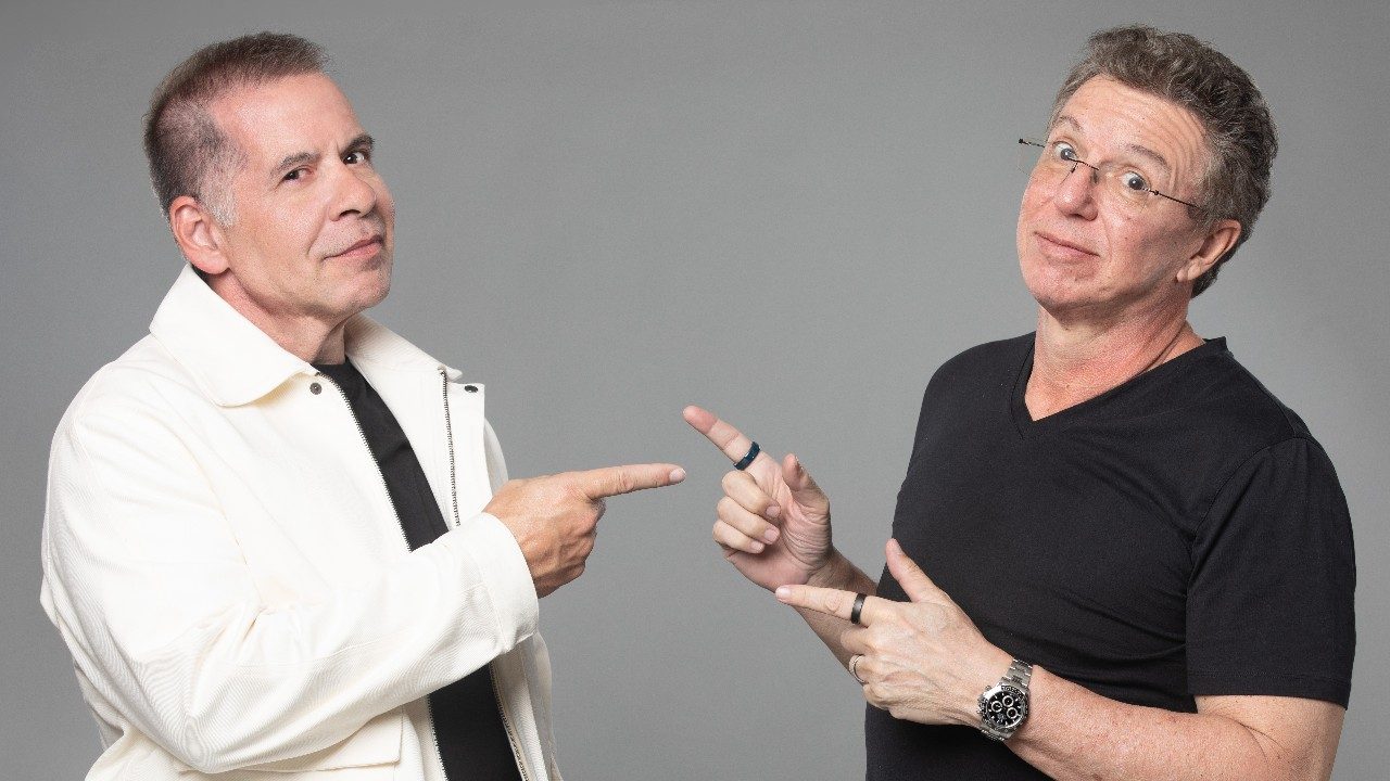Leandro Hassum e Boninho apontando um para o outro durante ensaio fotográfico em fundo cinza