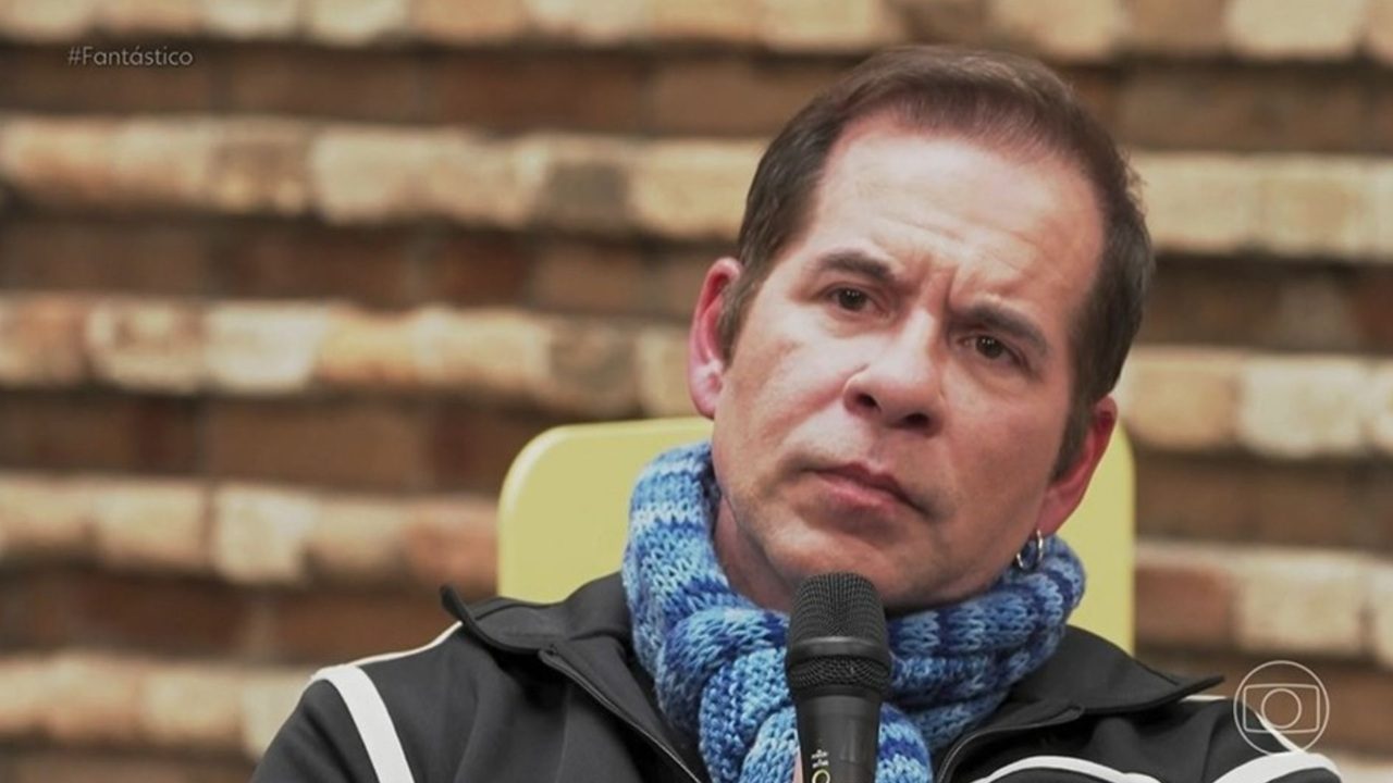 Homem com expressão séria segura um microfone durante entrevista, usando jaqueta preta com listras brancas e cachecol azul, com fundo de parede de tijolos desfocada e logotipo de programa de TV