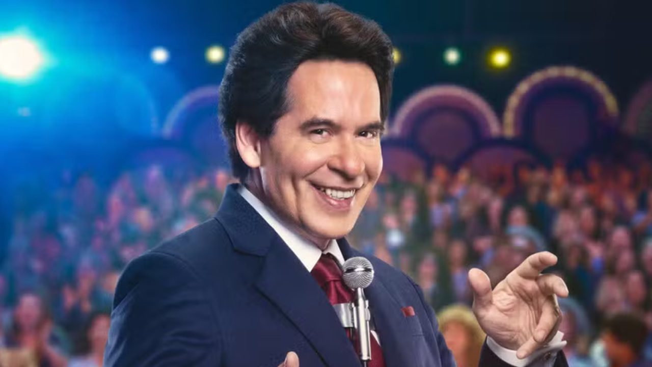 Homem sorridente de terno azul e gravata vermelha fala ao microfone em um palco iluminado, com plateia ao fundo. A expressão carismática e o ambiente colorido sugerem uma apresentação de programa de auditório ou show de variedades