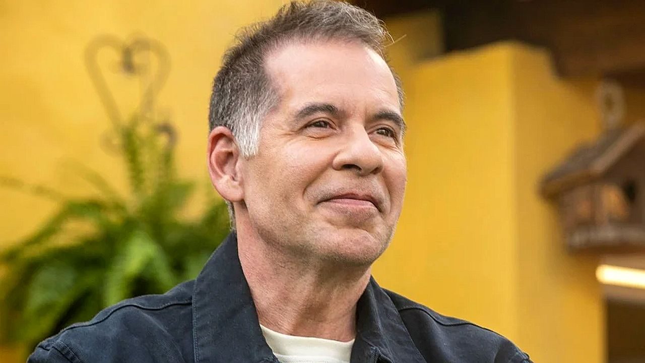 Homem sorri levemente em ambiente interno com parede amarela ao fundo em cena de programa de TV