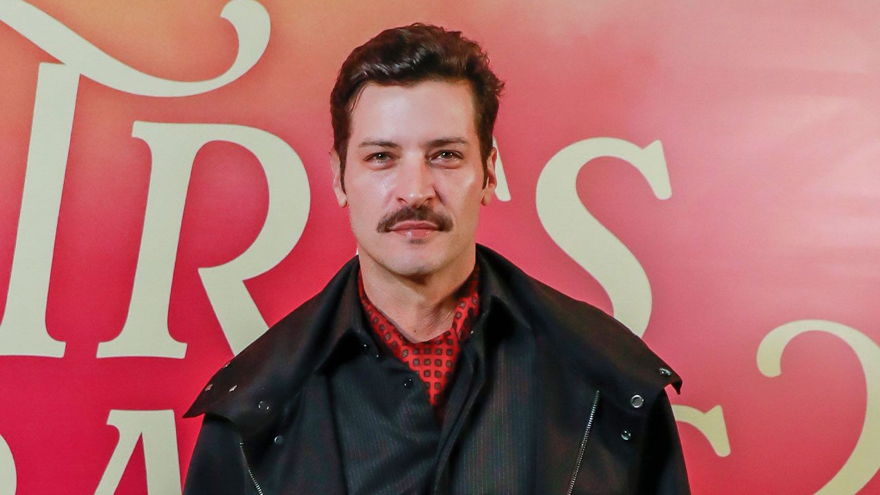 Leandro Lima, com bigode, camisa com lenço vermelho e jaqueta preta, posa para foto em evento de lançamento com painel vermelho ao fundo
