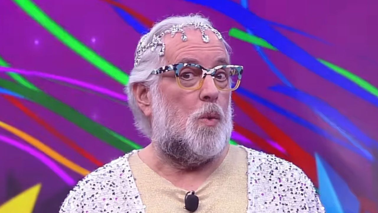 Leão Lobo olha para o lado com expressão de surpresa, o jornalista usa tiara de cristais, óculos coloridos e blusa de paetês prateados, fundo roxo