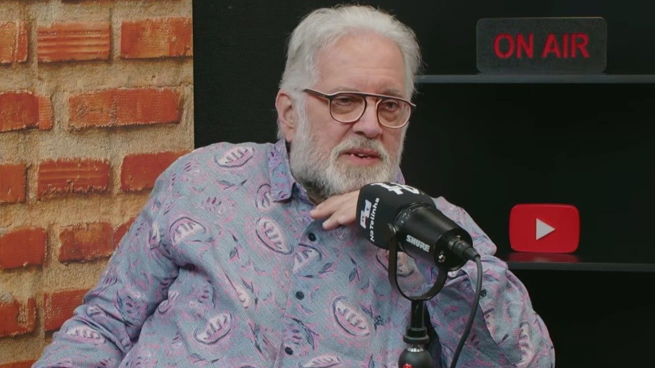 Leão Lobo de óculos e camisa estampada fala ao microfone em estúdio de podcast com placa On Air ao fundo