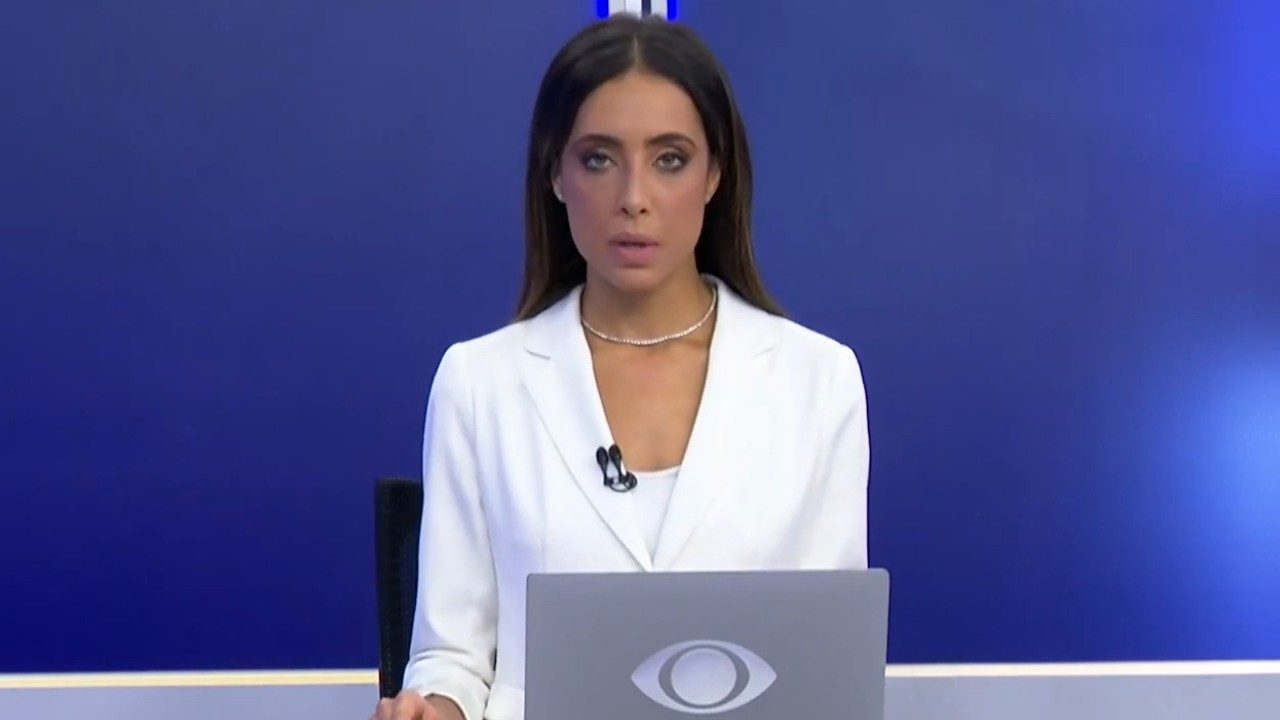 Lenny Leone de blazer branco apresenta telejornal atrás de bancada com notebook e fundo azul de estúdio