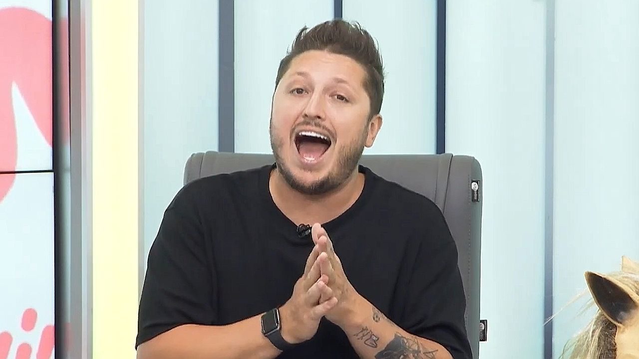 Apresentador de camiseta preta fala animado em programa de TV sentado em cadeira no estúdio