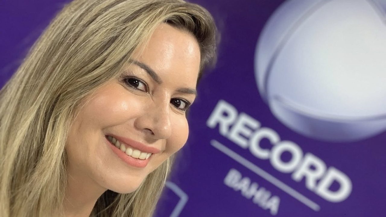 Mulher sorri em selfie tirada diante de um painel roxo com o logotipo da Record Bahia, indicando ambiente de emissora de TV