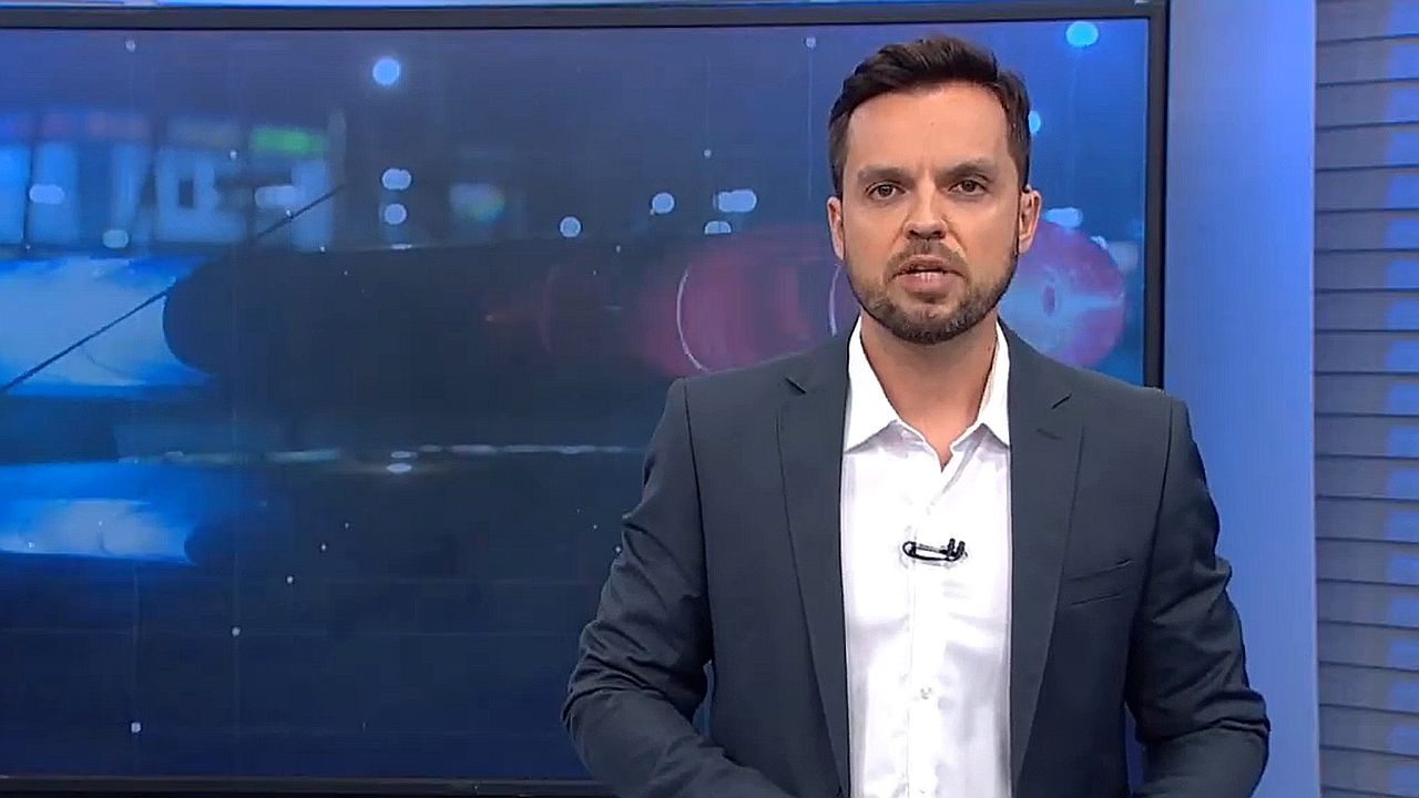 Apresentador de telejornal fala em estúdio com telão ao fundo exibindo imagem de viatura policial em destaque