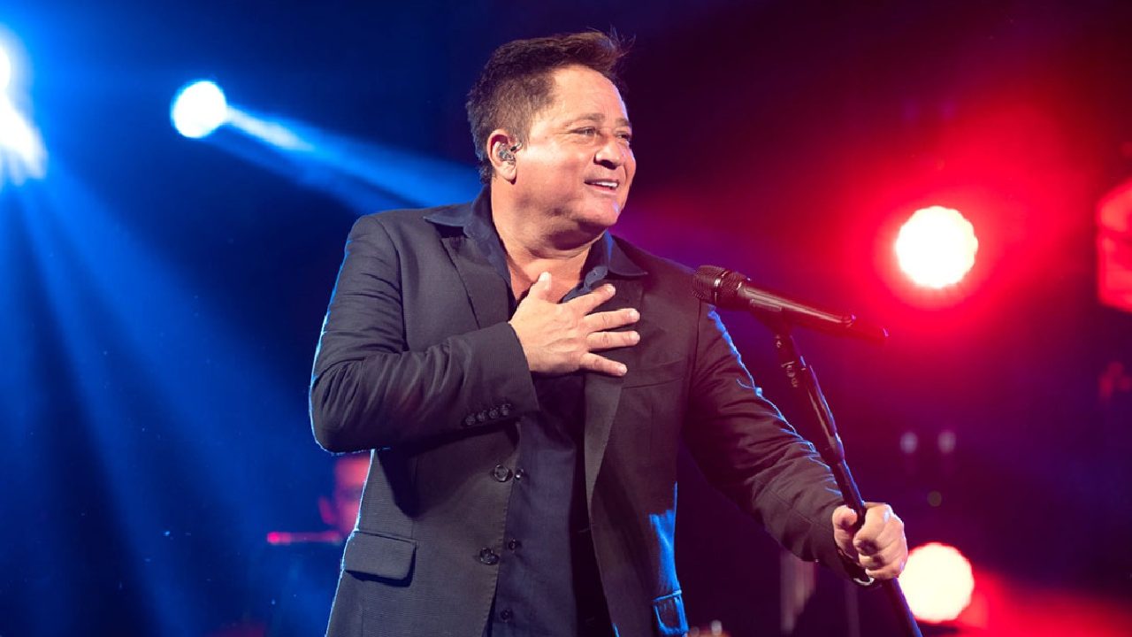 Cantor Leonardo se apresenta no palco, vestindo terno escuro e segurando o microfone enquanto sorri para o público, sob luzes coloridas de show