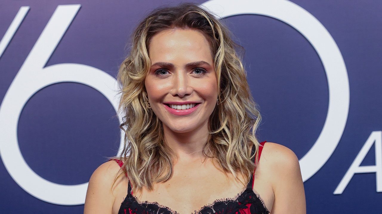 Letícia Colin com cabelo loiro ondulado e vestido preto com detalhes vermelhos em evento com fundo azul