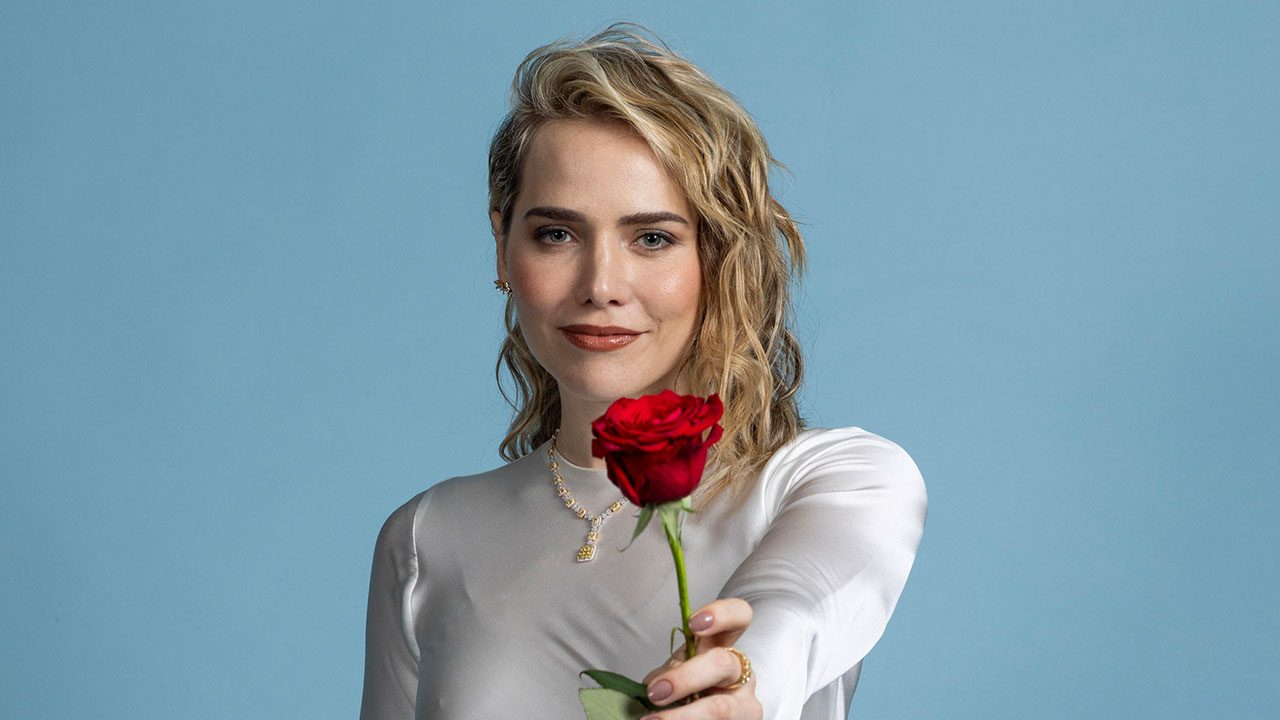 Leticia Colin com blusa branca e cabelo loiro ondulado segura uma rosa vermelha em direção à câmera com fundo azul claro