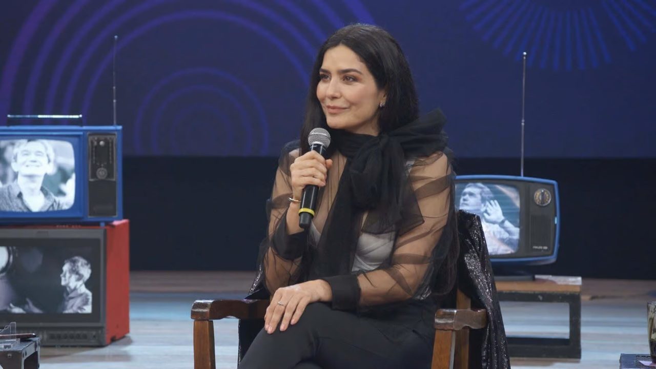 Mulher sentada em uma cadeira de madeira, segurando um microfone enquanto participa de uma conversa em um estúdio. Ela usa blusa preta translúcida com laço volumoso no pescoço e tem expressão serena. Ao fundo, aparecem televisores antigos exibindo imagens em preto e branco