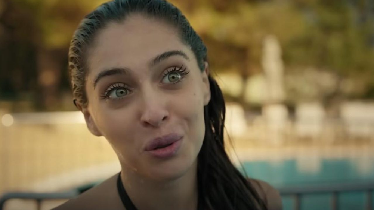 Mulher de cabelos molhados e expressão surpresa ou divertida aparece em close-up, com piscina e árvores desfocadas ao fundo em ambiente ensolarado