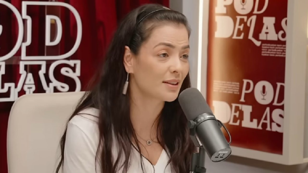 Li Martins durante participação no podcast PodDelas, falando ao microfone em estúdio decorado com cortina vermelha e painéis com o nome do programa ao fundo