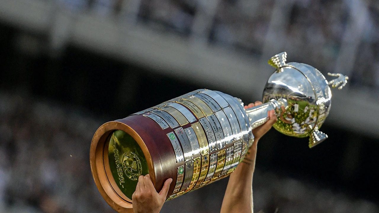 Mãos erguem o troféu da Copa Libertadores da América, uma taça prateada com base de madeira e placas douradas, diante de um estádio cheio de torcedores. A imagem simboliza celebração e conquista no futebol sul-americano