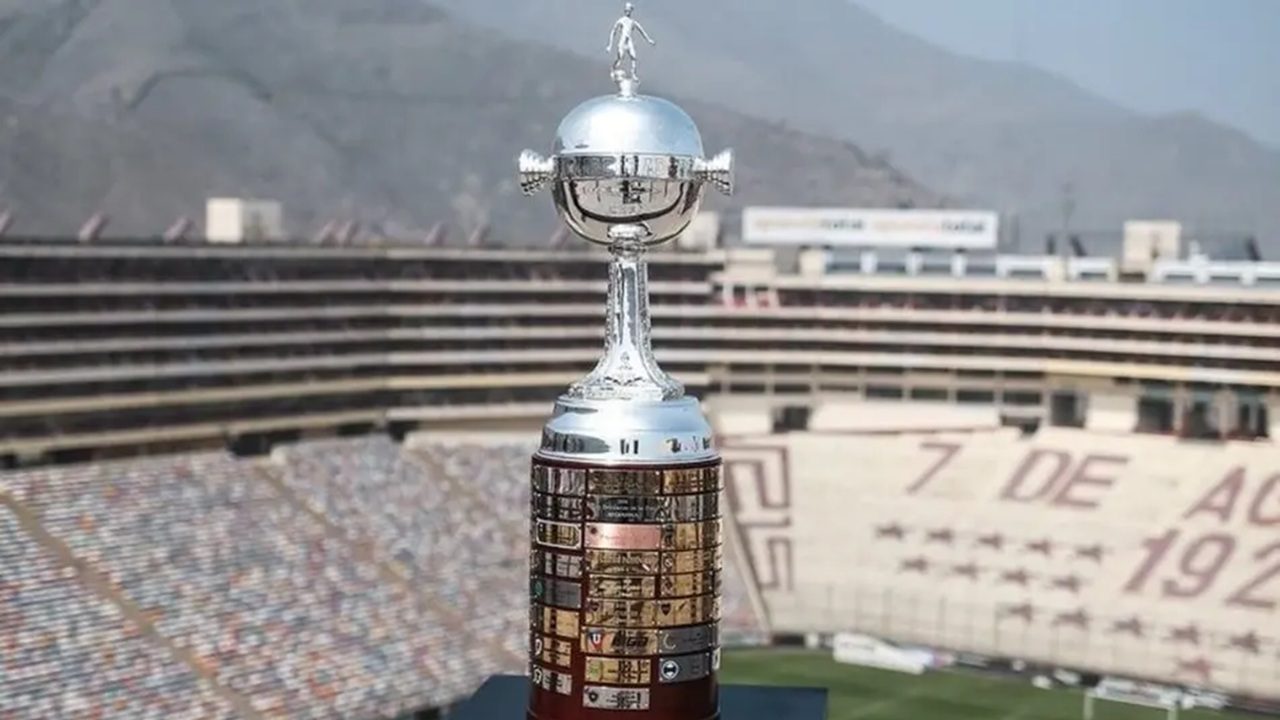 Troféu da Copa Libertadores da América em destaque, com acabamento prateado e base com placas douradas, posicionado no centro de um estádio vazio com arquibancadas e montanhas ao fundo.
