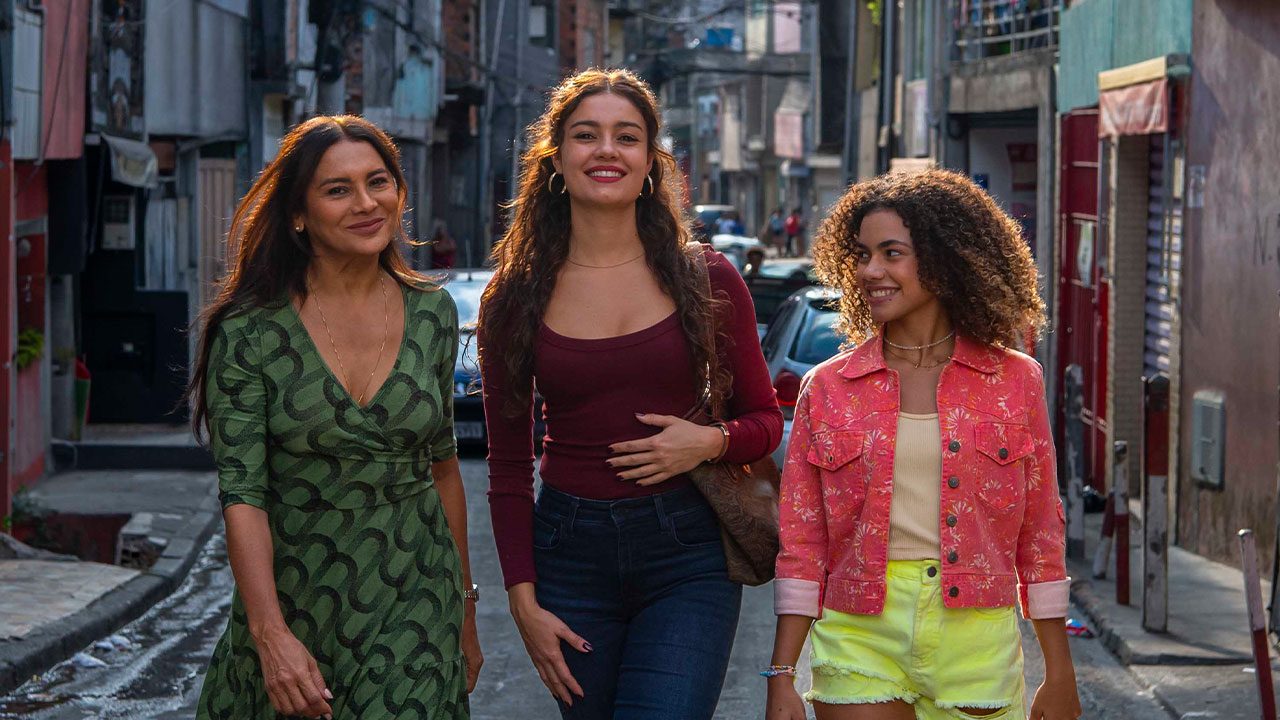 Lígia (Dira Paes), Gerluce (Sophie Charlotte) e Joélly (Alana Cabral) caminham sorrindo por uma rua de comunidade em cena da novela Três Graças