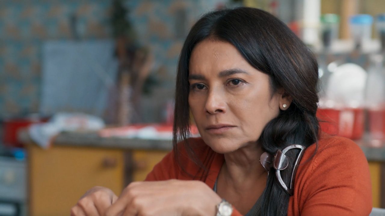 Dira Paes, com expressão séria e olhar concentrado, aparece sentada em ambiente doméstico com utensílios de cozinha ao fundo, usando cardigã laranja e prendedor grande no cabelo em cena da novela Três Graças, onde interpreta a personagem Lígia