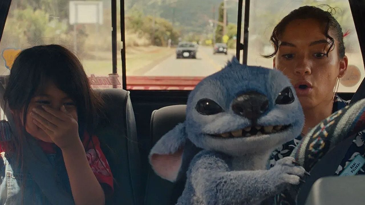 Dentro de um carro em movimento, duas meninas estão sentadas enquanto uma criatura azul animada, com aparência fofa e orelhas grandes, segura o volante e dirige. A jovem à direita parece surpresa, com a boca aberta, enquanto a menina à esquerda cobre o rosto, rindo ou reagindo à situação. Pela janela traseira, é possível ver a estrada e outros carros ao fundo, indicando que o veículo está em trânsito.