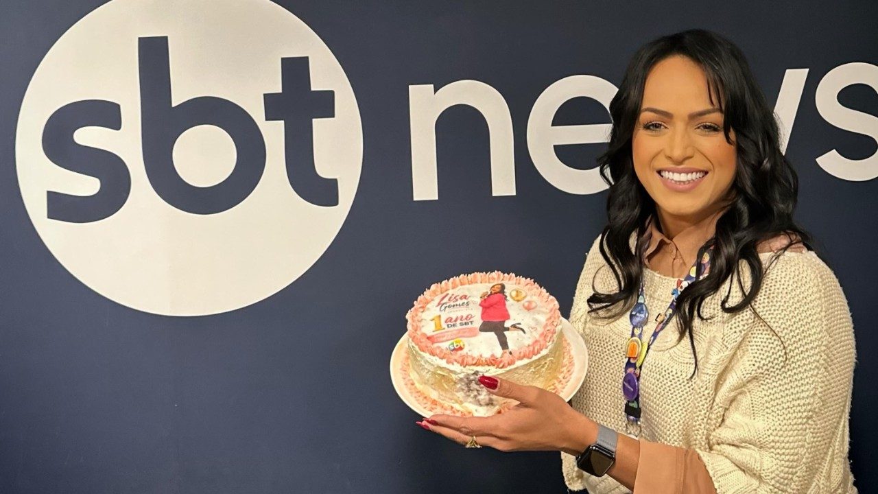 Lisa Gomes segura bolo comemorativo em frente ao logotipo do SBT News durante celebração de um ano na emissora