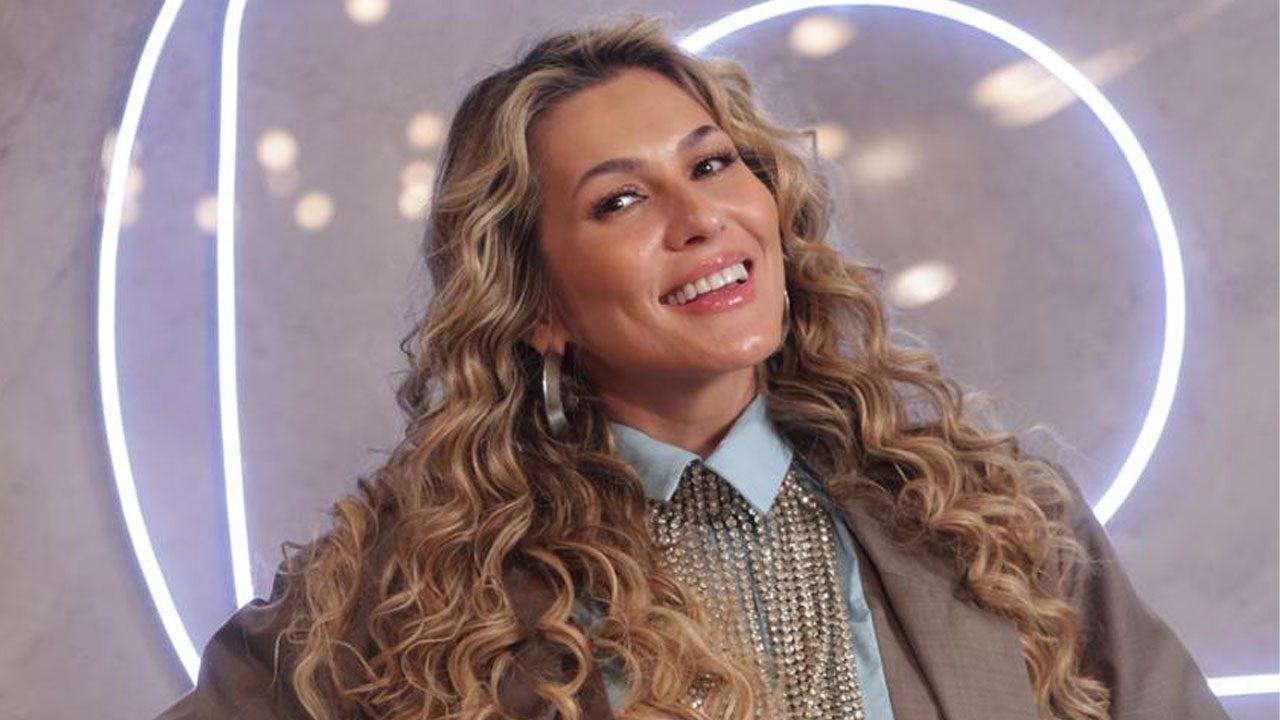 Lívia Andrade sorrindo, cabelo loiro cacheado, usando blazer, camisa azul e colar de strass em fundo iluminado