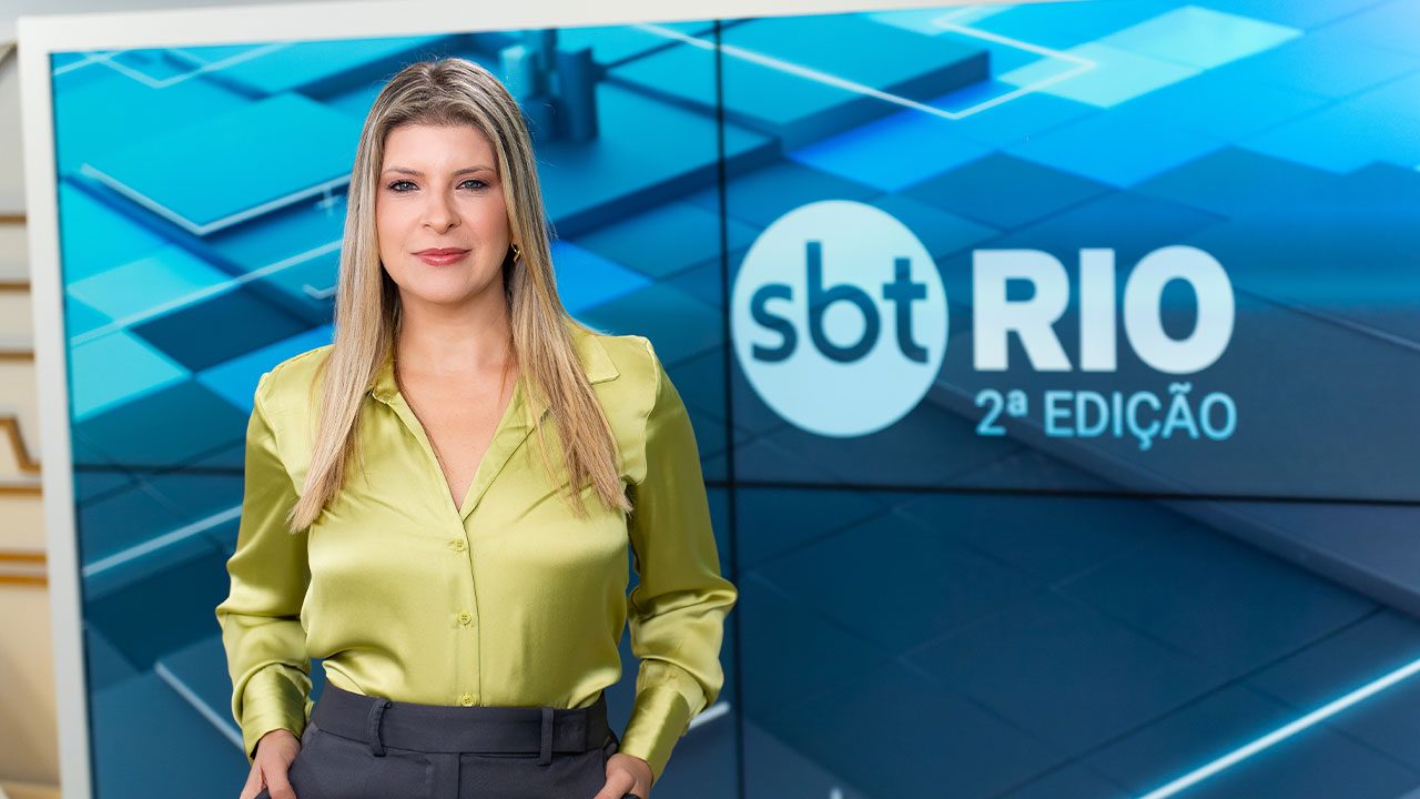 Lívia Mendonça em pé no estúdio do SBT Rio 2ª Edição com o telão do telejornal ao fundo